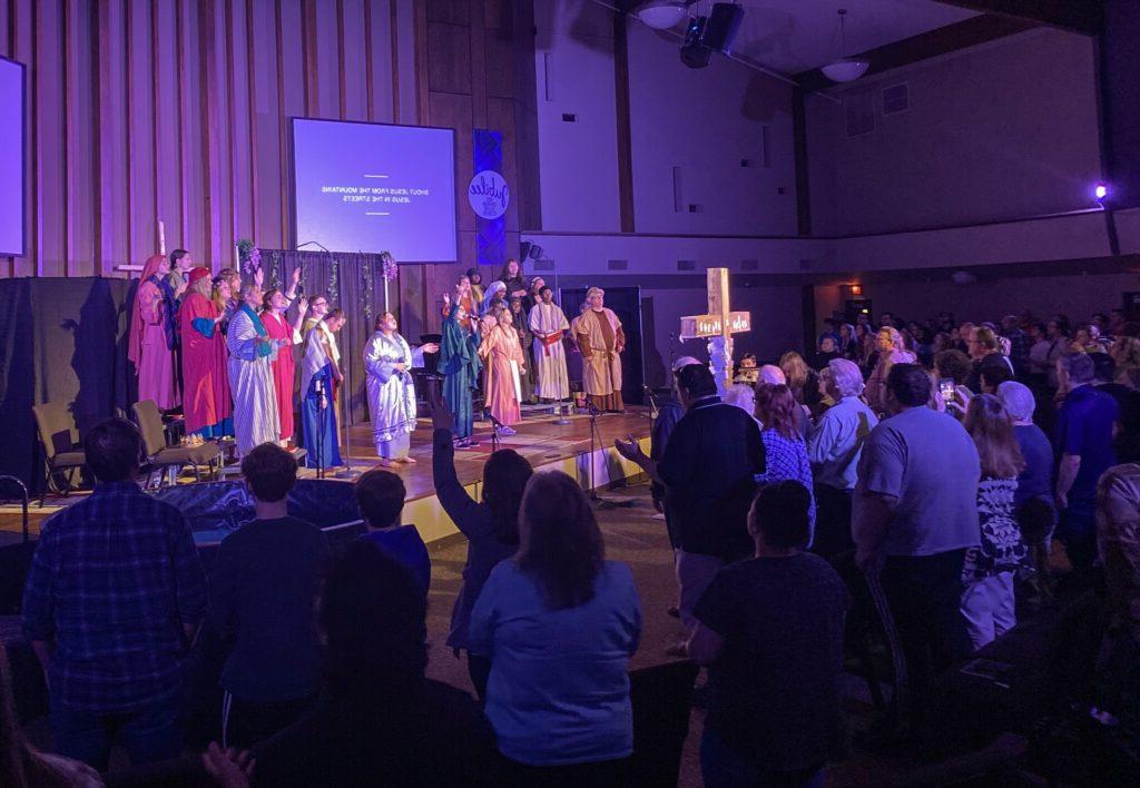 A group of people, dressed in costumes, perform on a stage in front of an audience inside a church. A wooden cross stands prominently at the forefront, partially obscuring stage view.